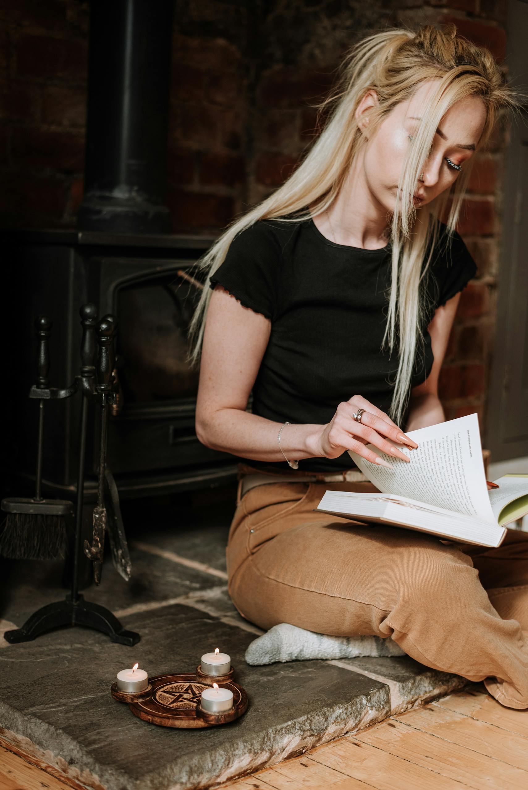 Serious blond female looking through pages of book during spiritual practice with candles and pentagram near fireplace